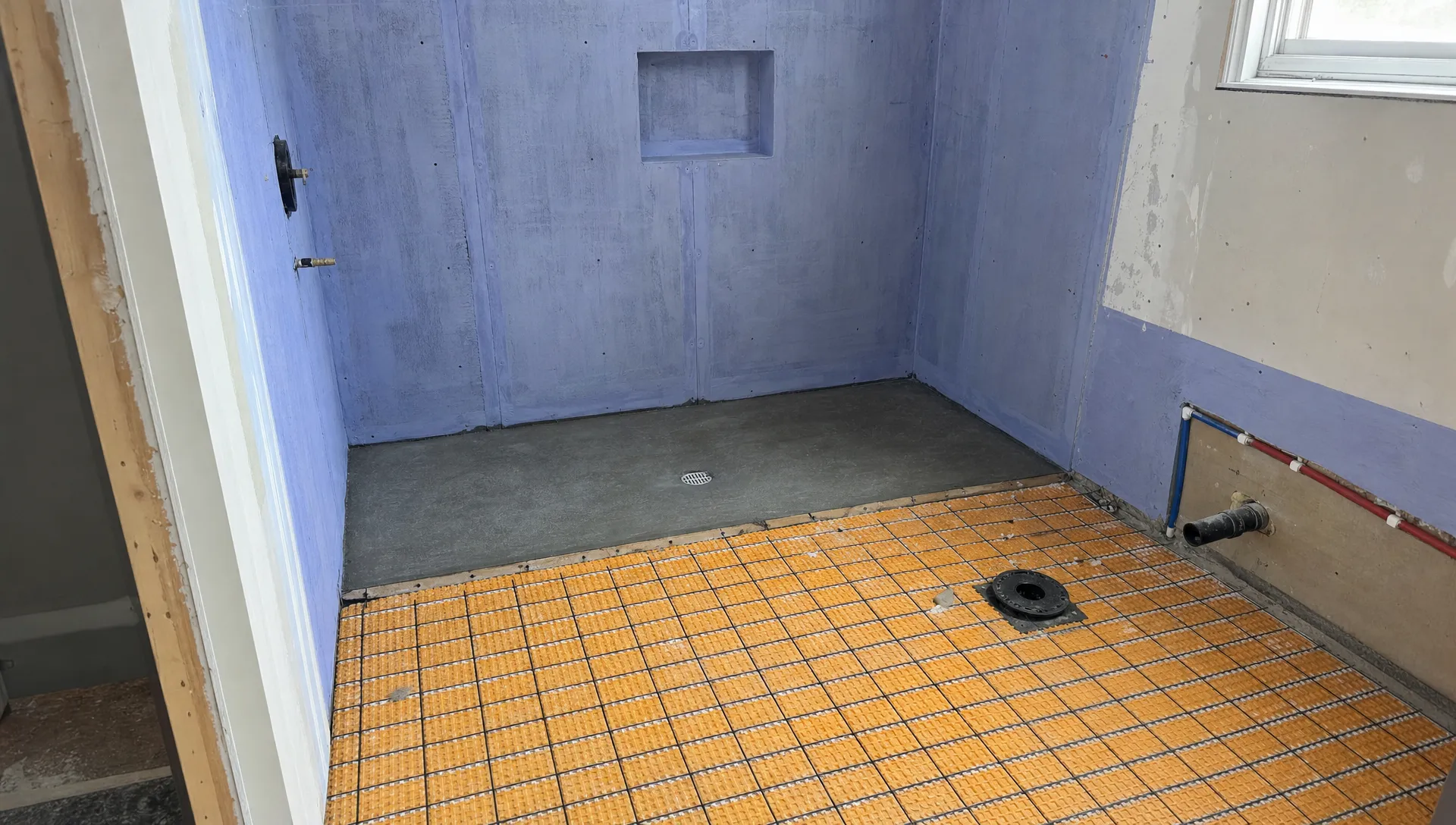 A partially renovated bathroom shows exposed plumbing lines, a shower drain, waterproofed walls, and floor preparation before tiling.
