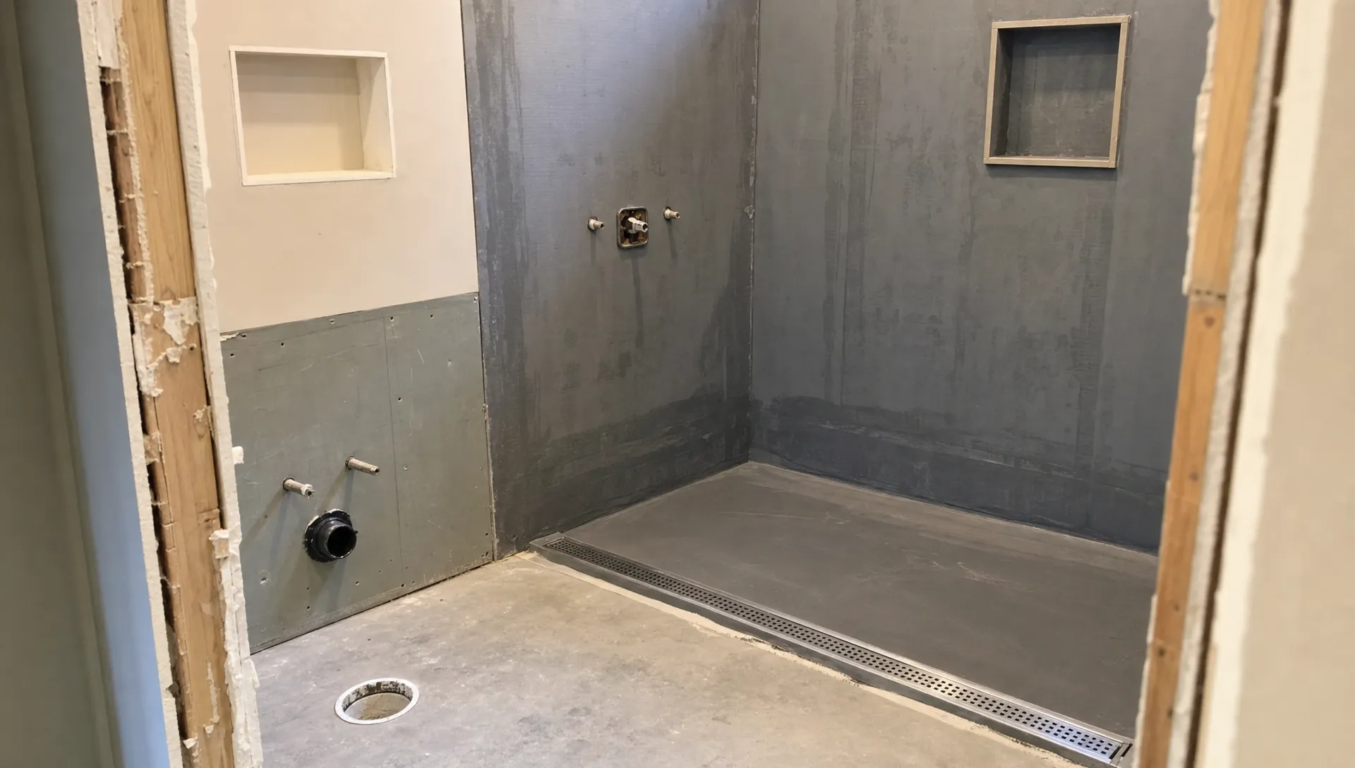 A mid-renovation bathroom shows exposed waterproofed walls, a prepared shower slope, plumbing outlets and a drain channel.